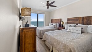 A hotel room with two beds, a ceiling fan, a large window with a view of the sea, and a wooden cabinet.
