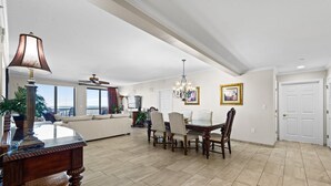 A spacious living area with a dining table, chairs, and a chandelier. There is a sofa, a TV, and a balcony visible.