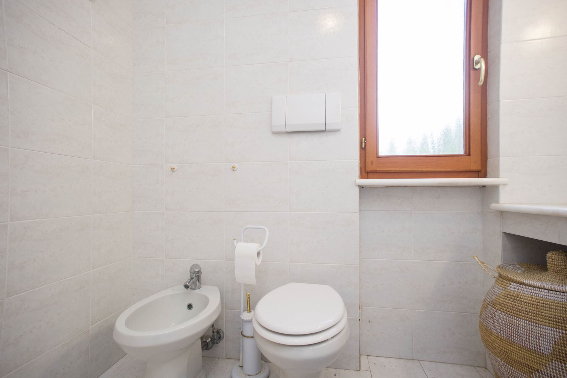 A small, clean bathroom with white tiles, a bidet, a toilet, and a window with a wooden frame.