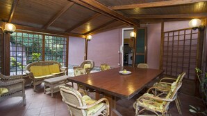 Una habitación amplia con una gran mesa de madera rodeada de sillas de mimbre, un sofá amarillo y vistas a la vegetación a través de amplios ventanales.