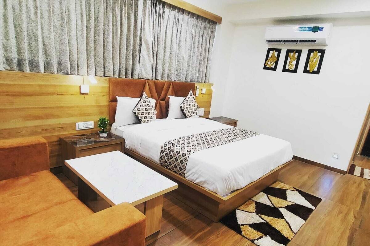 A modern hotel room with a wooden headboard, a large bed with patterned bedding, a bedside table, a small table, and a patterned rug.