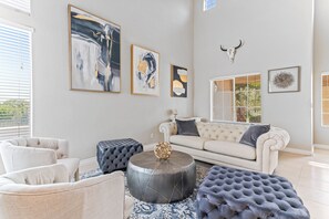 A living room with a white sofa, two ottomans, and a coffee table. There are three framed artworks on the wall and a bull skull decoration. - Amazing 5 BDR, Resort Style Pool w/Heater and Games. (Henderson)