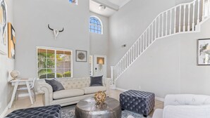 Un salón amplio con una escalera blanca, una mesa de centro redonda y un objeto decorativo, y un ventanal con persianas.