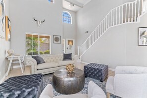 A spacious living room with a white staircase, a round coffee table with a decorative object, and a large window with blinds. - Amazing 5 BDR, Resort Style Pool w/Heater and Games. (Henderson)