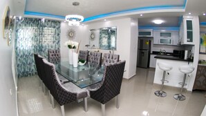 A dining area with a glass table, cushioned chairs, a chandelier, and a kitchen in the background. - ✨ Luxurious 3-Bedroom Apartment with Pool & 24/7 security  (Santiago De Los Caballeros)