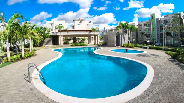 A swimming pool with a curved design, surrounded by a paved walkway and palm trees. - ✨ Luxurious 3-Bedroom Apartment with Pool & 24/7 security  (Santiago De Los Caballeros)