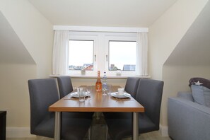 Un coin repas avec une table dressée pour deux personnes, deux chaises, une bouteille, des verres et une tasse. Il y a une fenêtre avec des rideaux et une vue sur les bâtiments à l’extérieur.