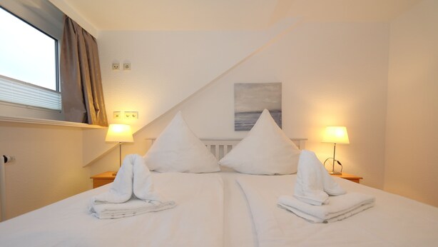 Une chambre d’hôtel avec deux lits, chacun étant agrémenté d’une serviette pliée et d’un oreiller. On y trouve deux lampes de chevet et une fenêtre avec des rideaux.