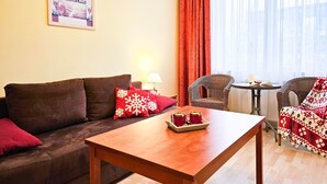 Ein gemütliches Wohnzimmer mit einem braunen Sofa, roten Kissen, einem Holztisch mit zwei roten Tassen, einem Korbsessel und Blick ins Freie durch transparente Vorhänge.