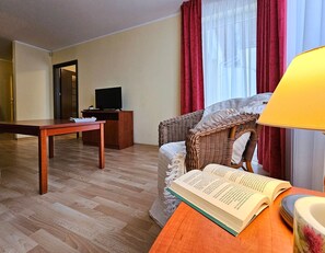 Une chambre d’hôtel avec un plancher en bois, une table à manger, un canapé en osier, un téléviseur posé sur un meuble et une lampe.