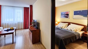 A hotel room with a wooden floor, a bed with a quilt, a desk with a TV, a window with curtains, and a wall with two framed pictures. - Naut 310 - Nautilius (Swinoujscie)