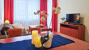 A living room with a sofa, dining table, and TV. There are red curtains, a vase with flowers, and a lamp. - Naut 310 - Nautilius (Swinoujscie)