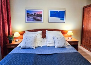 Un lit bien fait, recouvert d’une couette bleue, avec deux oreillers et une table de chevet de chaque côté, sur laquelle se trouve une lampe. Deux tableaux encadrés sont accrochés au mur, au-dessus du lit.