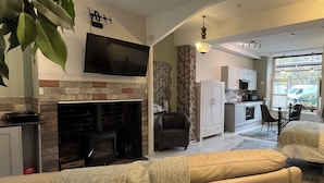 Sala de estar con chimenea, sofá y televisor. Se observa una zona de cocina con armarios y una mesa de comedor.