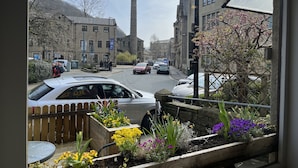 Vista de la calle con coches aparcados en la acera, una chimenea alta al fondo y diversos tipos de flores en macetas al frente.