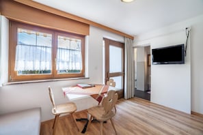 A room with a dining table, chairs, a TV mounted on the wall, and large windows with curtains.
