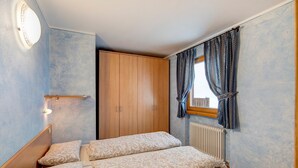 A bedroom with a wooden wardrobe, a bed with patterned bedding, a window with curtains, and a wall-mounted light fixture. - Apartment "Piz Filone" with Mountain View, Wi-Fi (Livigno)