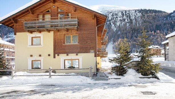 A wooden chalet with a balcony, a stone entrance, and a view of snow-covered mountains. - Apartment "Piz Filone" with Mountain View, Wi-Fi (Livigno)
