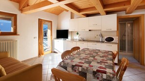 Une cuisine avec des poutres en bois, des meubles blancs, une table à manger avec des chaises, un téléviseur et un espace douche.