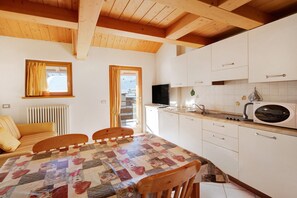 A room with a wooden ceiling, a quilted table, a sofa, a kitchen with white cabinets, a microwave, and a TV.