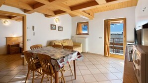 Une pièce chaleureuse avec un plafond en bois, une table à manger avec des chaises, un canapé, un radiateur et une vue sur l’extérieur à travers une fenêtre avec des rideaux.