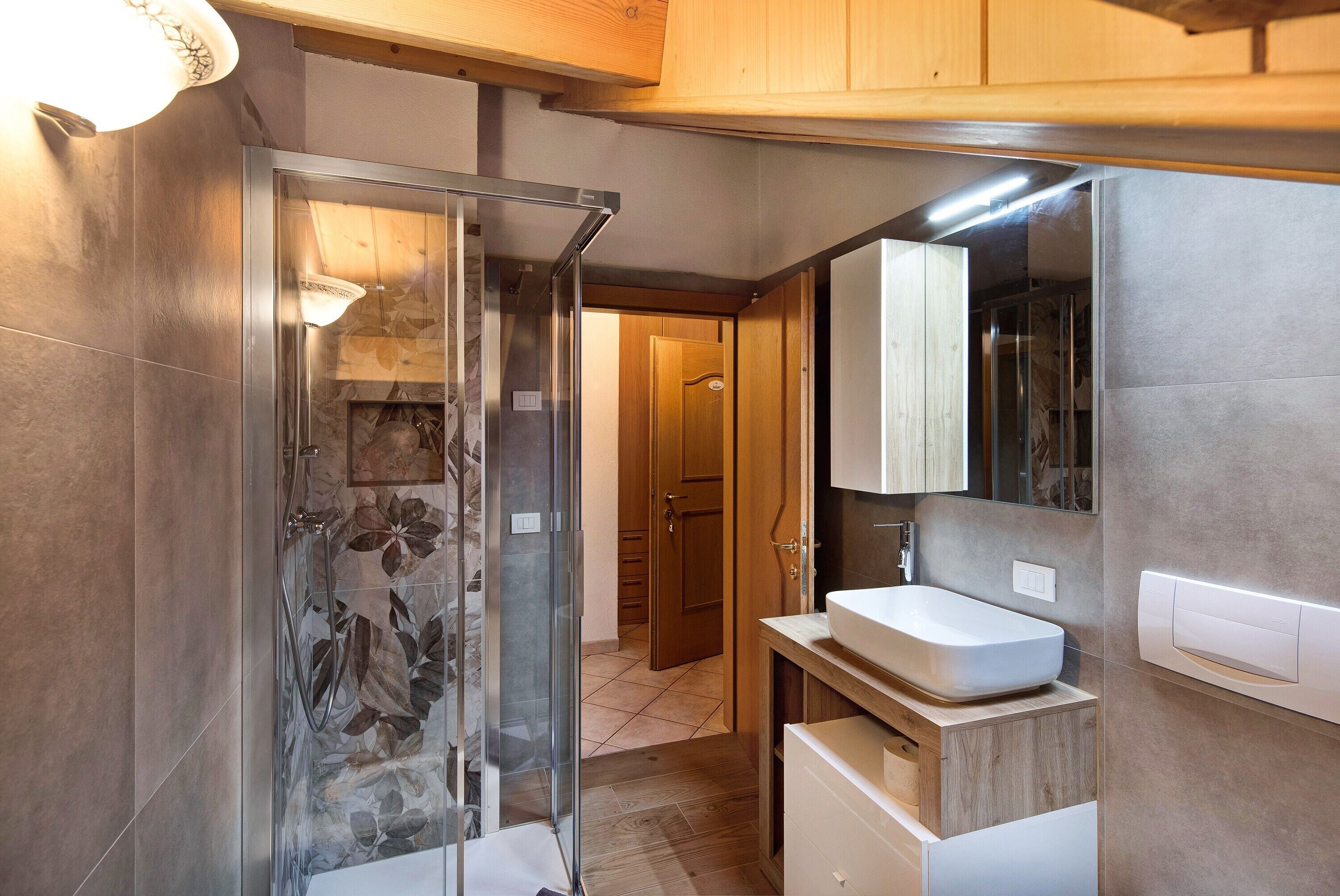 A bathroom with a glass shower enclosure, a white sink, and wooden cabinets.
