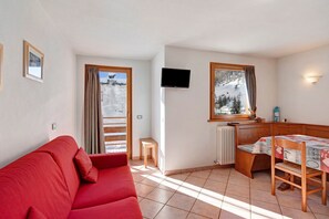 A room with a red sofa, a wooden table with a tablecloth, a TV mounted on the wall, and a window with a view of a snowy landscape. - 'Appartamento Vago' with Mountain View, Wi-Fi, Balcony & Garden (Livigno)
