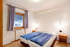 A bedroom with a bed, two bedside tables, a window with curtains, a radiator, and a wall-mounted light fixture.