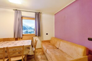 A room with a wooden table, chairs, a sofa, a window with curtains, and a view of mountains.