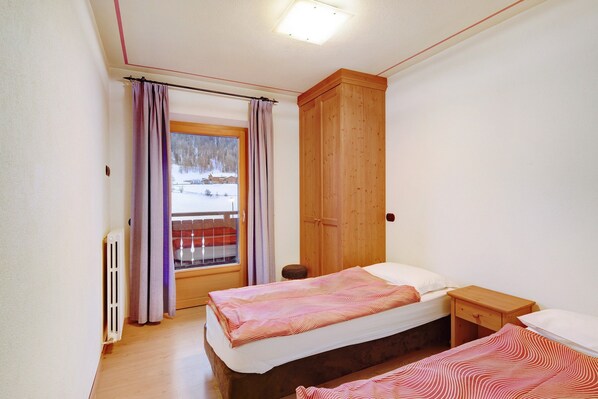 A bedroom with two beds, a wooden wardrobe, a window with curtains, and a radiator.