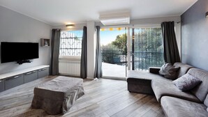 A modern living room with a flat-screen TV, grey sofa, ottoman, and a large sliding glass door leading to a balcony.