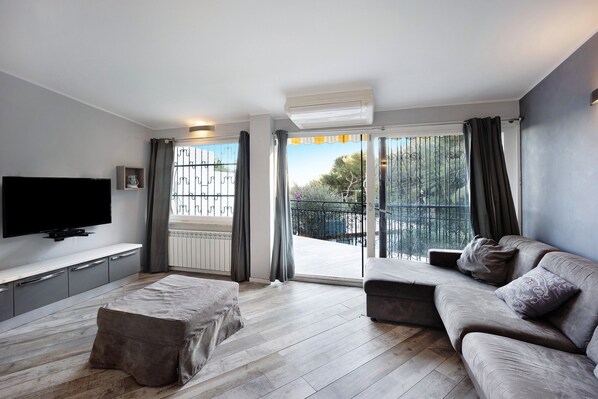 A modern living room with a flat-screen TV, grey sofa, ottoman, and a large sliding glass door leading to a balcony.