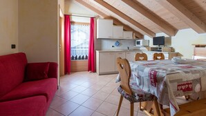 A cozy room with a red sofa, a wooden dining table with chairs, a kitchen area with cabinets and a microwave, and a window with curtains. - Apartment Stefano 2 with Mountain View, Wi-Fi & Garden (Livigno)