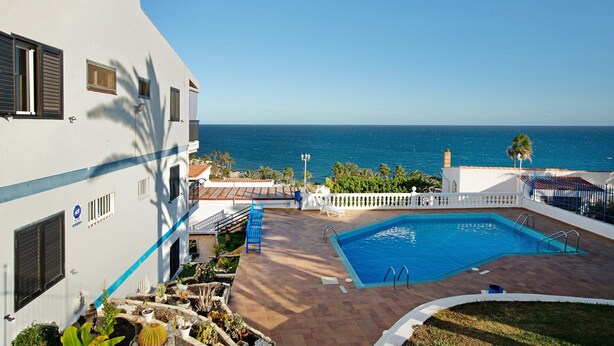 Área da piscina com vista para o mar, um edifício branco com portadas e a borda da piscina azul.