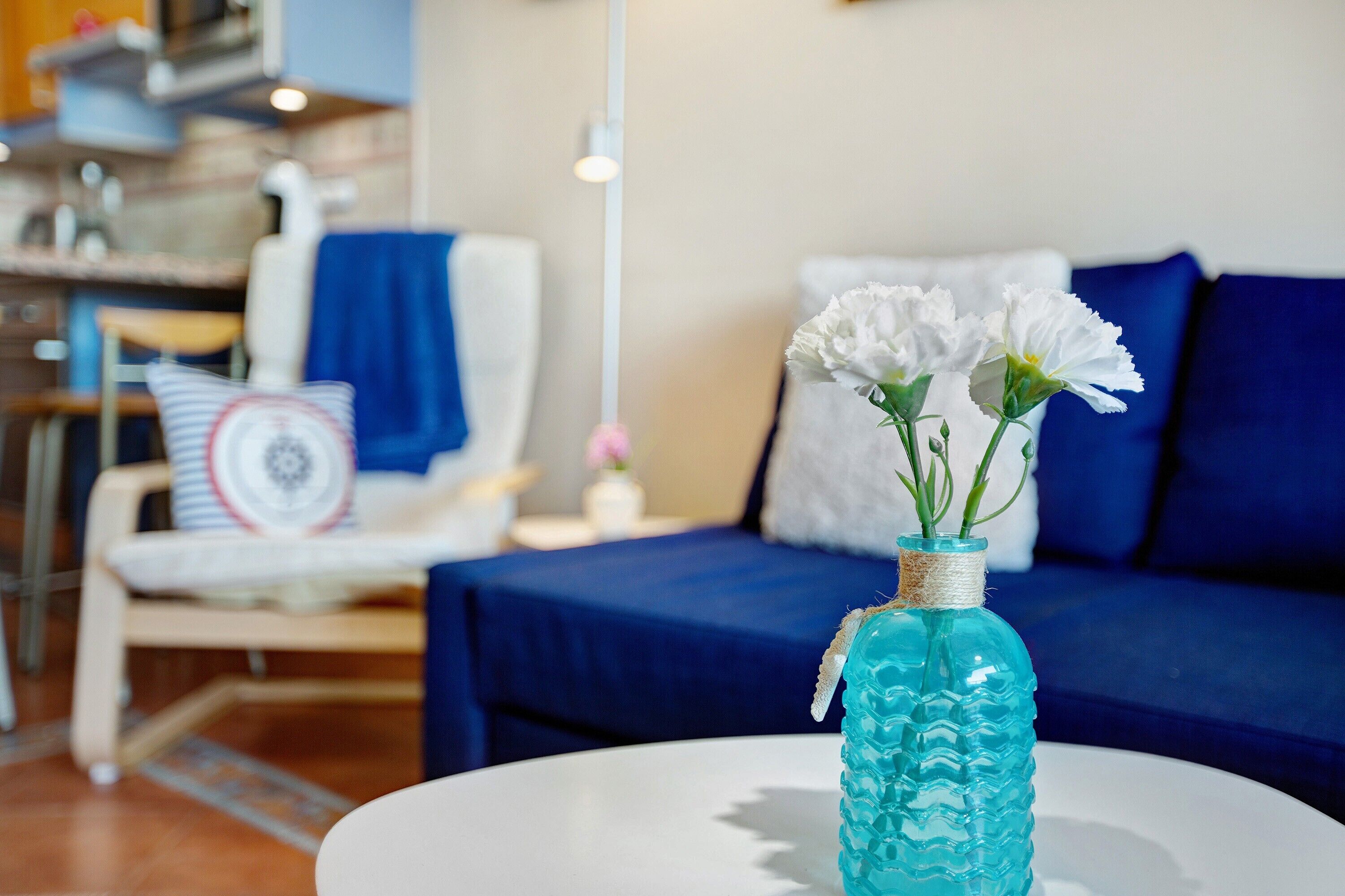 En stue med en blå sofa, en hvid stol og et rundt bord med en blå vase med hvide blomster.