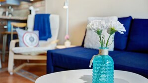 Uma sala de estar com um sofá azul, uma cadeira branca e uma mesa redonda com um vaso azul contendo flores brancas.