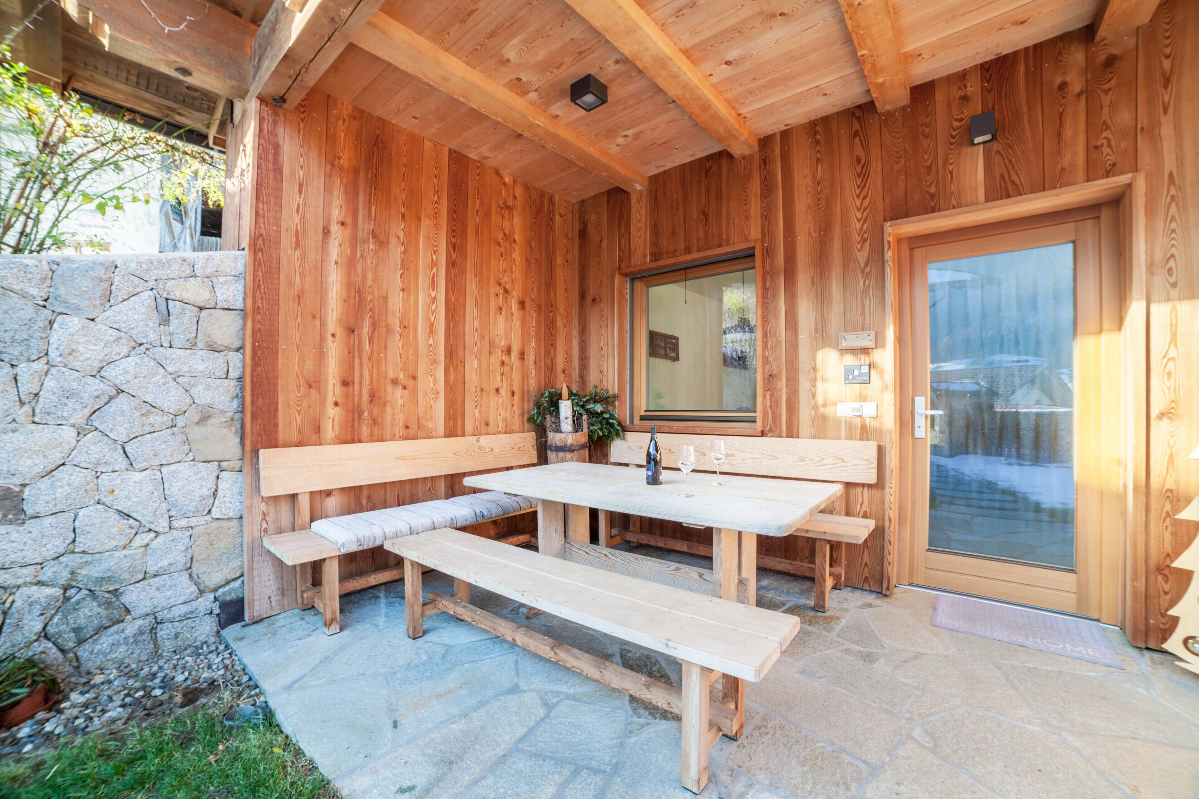 A wooden exterior with stone walls, a wooden bench, and a table with a bottle and glasses.