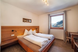Une chambre à coucher avec une tête de lit en bois, un lit recouvert de draps blancs, un petit bureau et une vue sur les montagnes par la fenêtre.