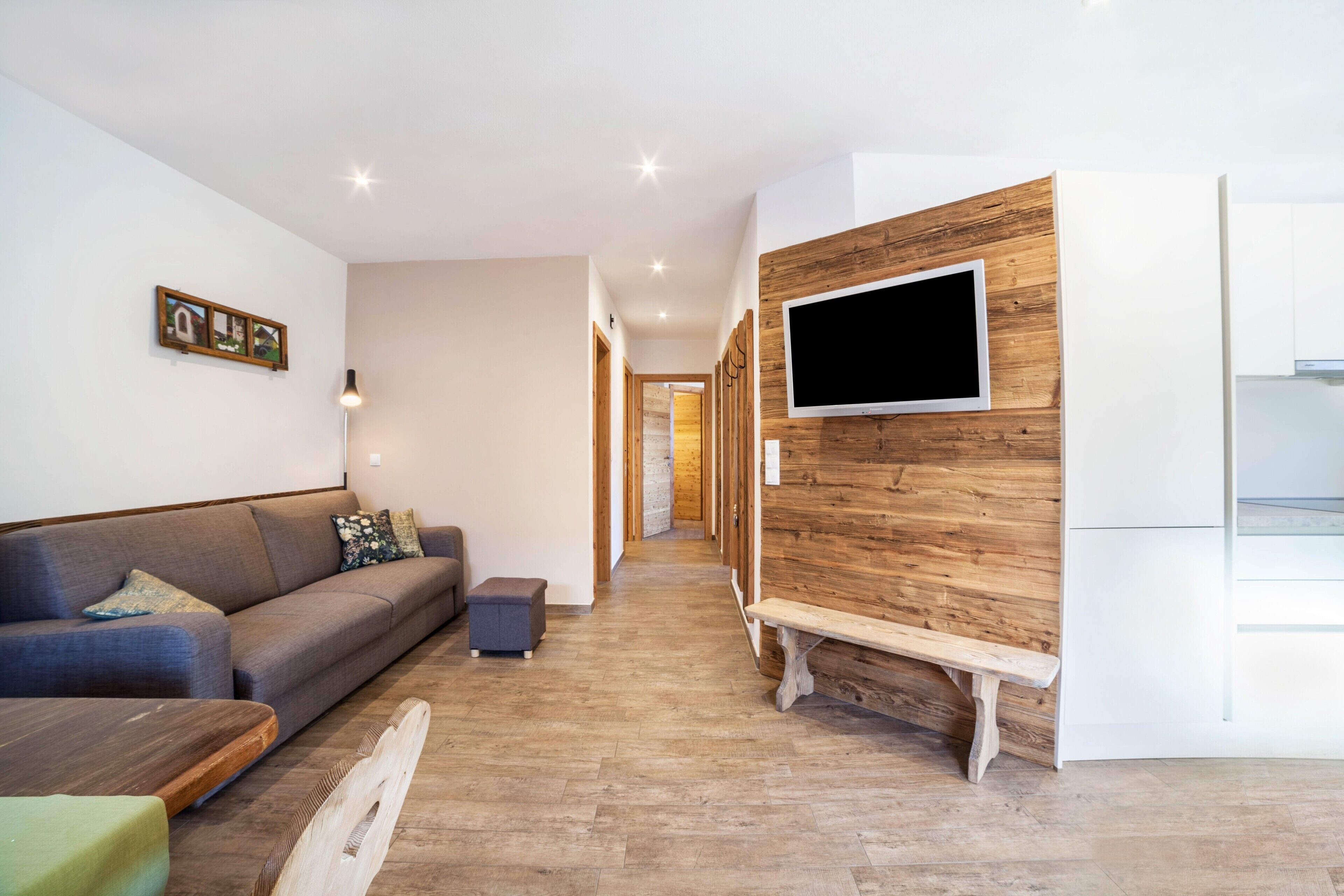 A modern living room with a sofa, a bench, a TV mounted on a wooden wall, and a hallway leading to another room.