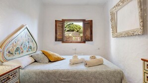 A bedroom with a bed, a window with an open shutter, a wall-mounted picture frame, and a bedside table.