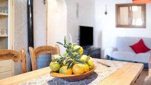 A dining table with a bowl of lemons and oranges, wooden chairs, a television, and a white sofa with a red pillow in a living room.