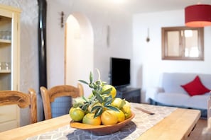A dining table with a bowl of lemons and oranges, wooden chairs, a television, and a white sofa with a red pillow in a living room. - Holiday Home Casa Inma with Pool, Garden and Wi-Fi (Vejer de la Frontera)