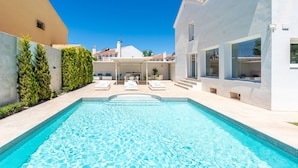 Piscina com espreguiçadeiras, área de estar coberta e um edifício branco com grandes janelas.