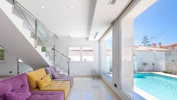 Interior moderno com escada de vidro, vista para piscina e sofá com almofadas.