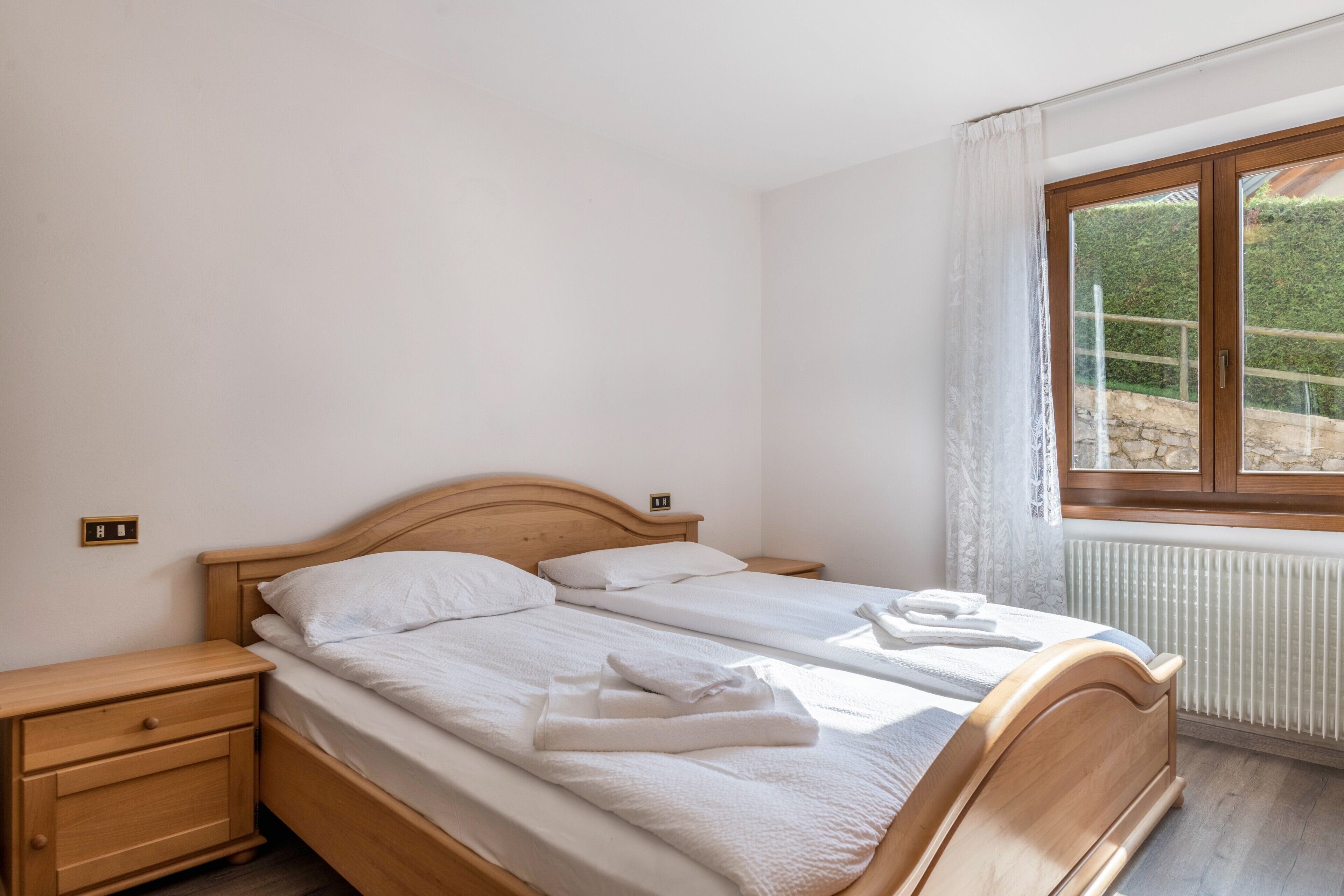 Un dormitorio con dos camas individuales, una mesita de noche de madera, una ventana que ofrece vistas a zonas verdes y un radiador.