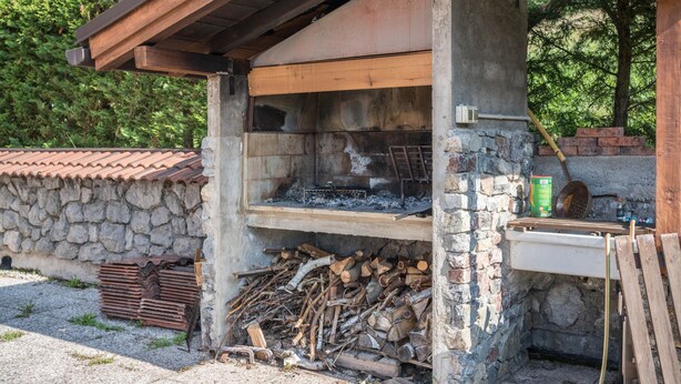 Un horno de piedra al aire libre con chimenea, una pila de leña y un recipiente metálico.