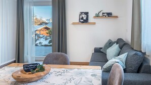 Una sala de estar moderna con un sofá gris, una mesa de centro de madera con un letrero de bienvenida y vistas a un paisaje nevado a través de un ventanal grande.