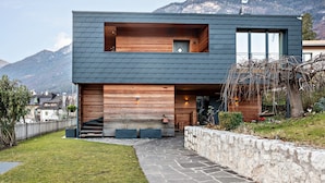 A modern two-story house with a stone pathway leading to the entrance, a wooden deck, and a view of mountains in the background. - Apartment 'Villa KaTo' with Lake View, Garden & Terrace (Kaltern an der Weinstraße)