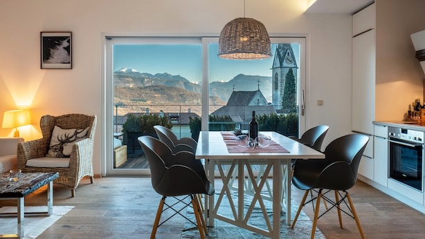 A modern living room with a dining area, a wicker chair, a table with chairs, a lamp, and a view of mountains and a church through large windows. - Apartment 'Villa KaTo' with Lake View, Garden & Terrace (Kaltern an der Weinstraße)