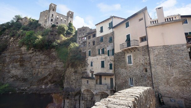 Un pueblo costero con edificios históricos de piedra, un castillo imponente en un acantilado y un puente de piedra.